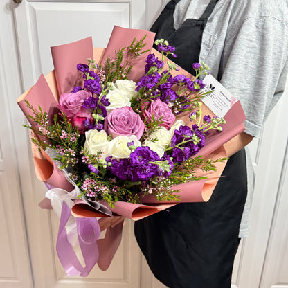 Vibrant bouquet of white, lavender, and purple flowers wrapped in pink paper.