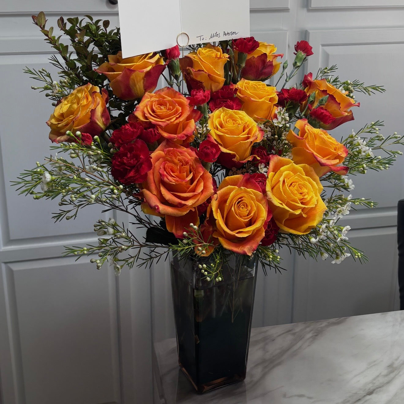 Vibrant bouquet of orange and yellow roses with red carnations in a dark vase.