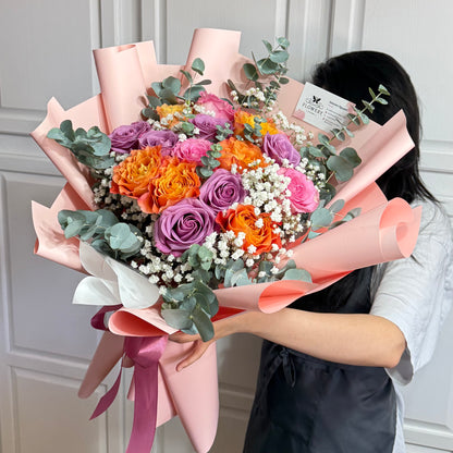 A person holds a vibrant bouquet of orange, purple, and pink roses wrapped in pink paper.