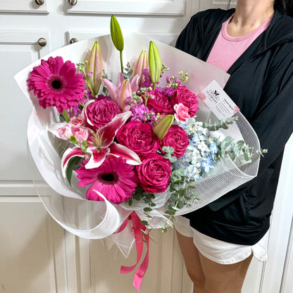 Vibrant pink lilies, roses, and daisies bouquet wrapped in white mesh.