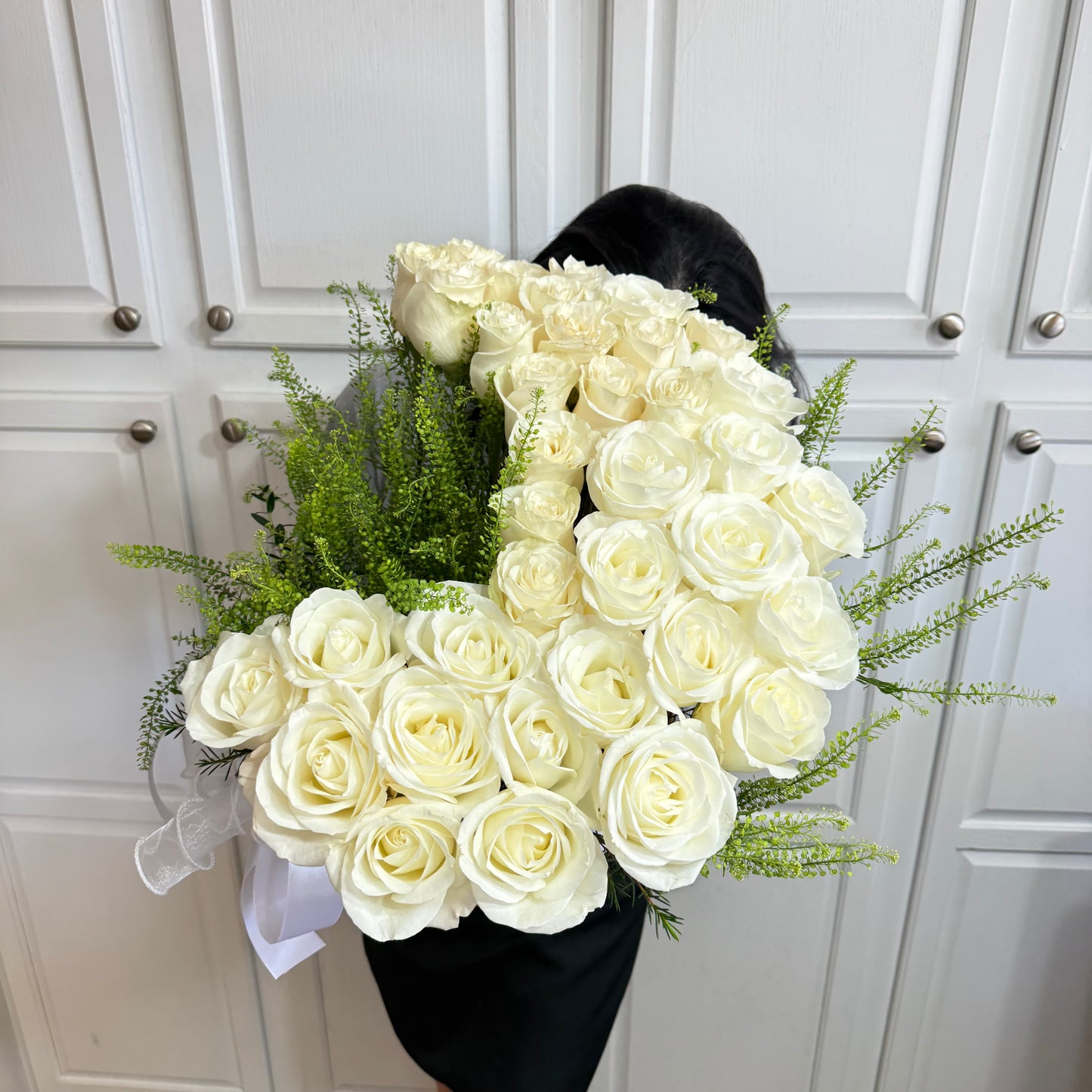 Person holding a large, full bouquet of white roses with green foliage.