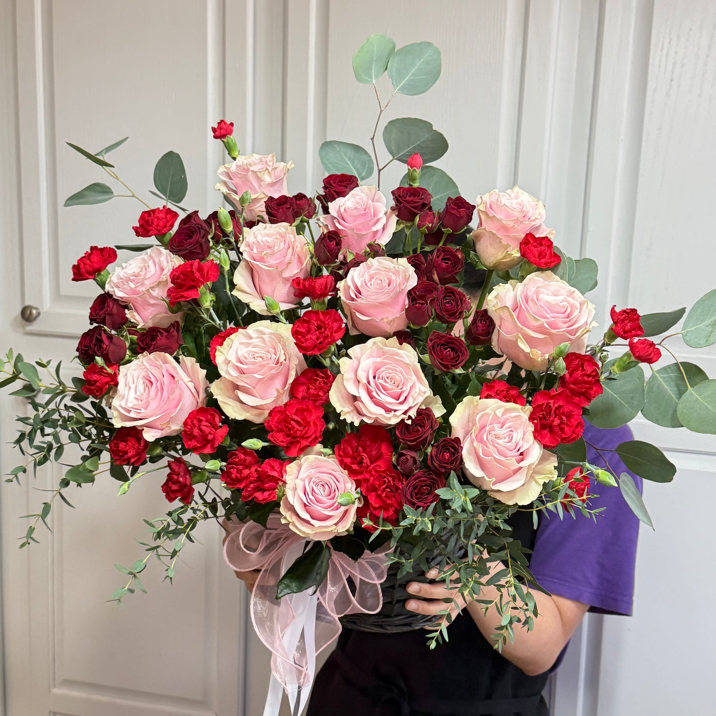 Large bouquet of pink roses, red carnations, and eucalyptus in a dark basket.