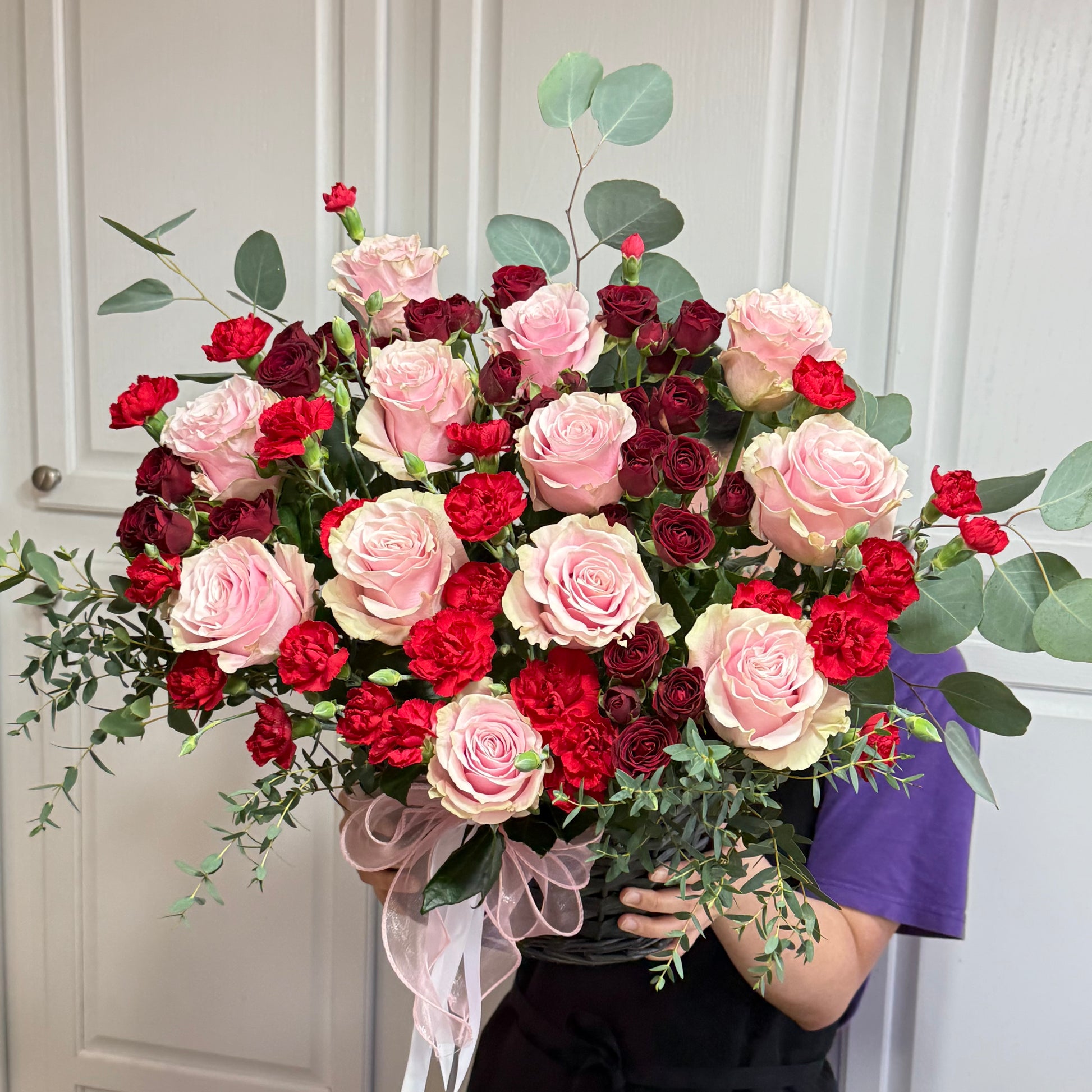 Large bouquet of pink roses, red carnations, and eucalyptus in a dark basket.