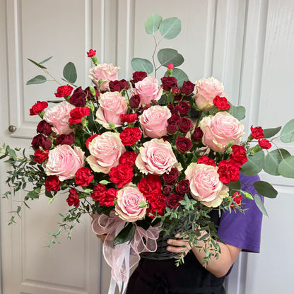 Large bouquet of pink roses, red carnations, and eucalyptus in a dark basket.