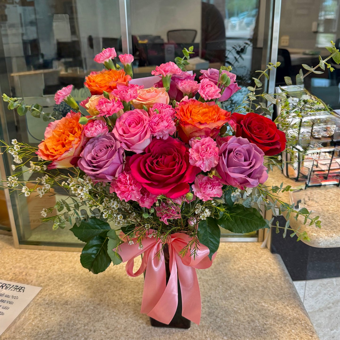 Vibrant pink, orange, red, and purple flower bouquet with a pink ribbon on a dark vase.