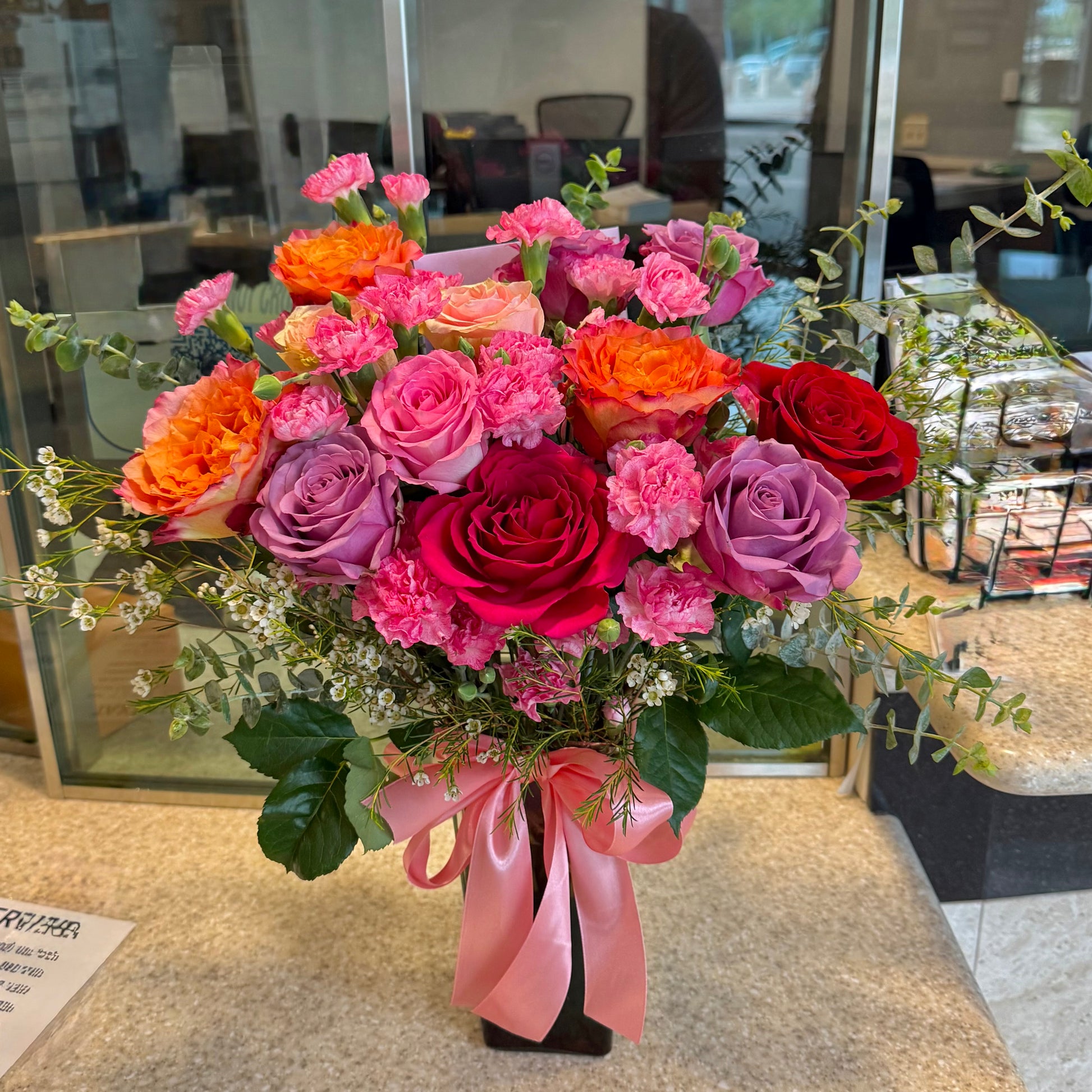 Vibrant pink, orange, red, and purple flower bouquet with a pink ribbon on a dark vase.