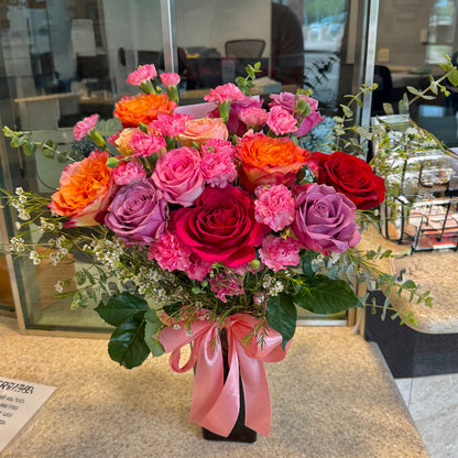 Vibrant pink, orange, red, and purple flower bouquet with a pink ribbon on a dark vase.