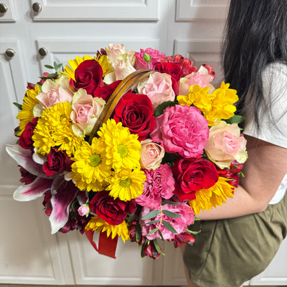 A person holds a large, colorful flower arrangement in a basket with roses and lilies.