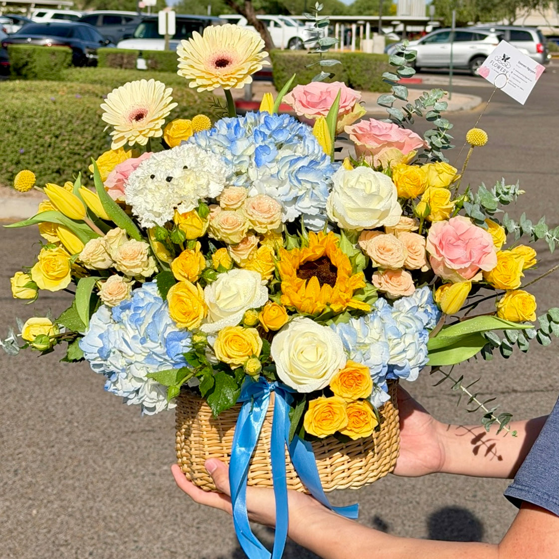 Hands holding a wicker basket of vibrant yellow, blue, and pink spring flowers.