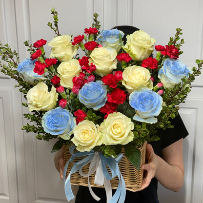 A large wicker basket filled with yellow, blue, and red roses and carnations.