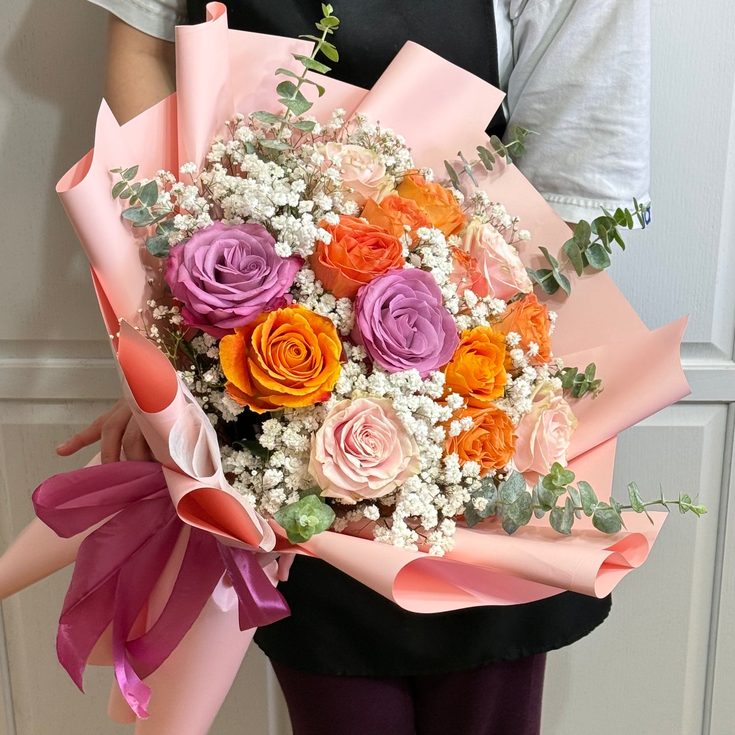 A person holds a vibrant bouquet of orange, purple, and pink roses in pink wrapping.