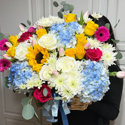 Vibrant bouquet of white, yellow, blue, and pink flowers in a woven basket.