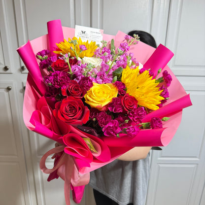 Vibrant bouquet of red roses, yellow blooms, and purple flowers in pink wrap.