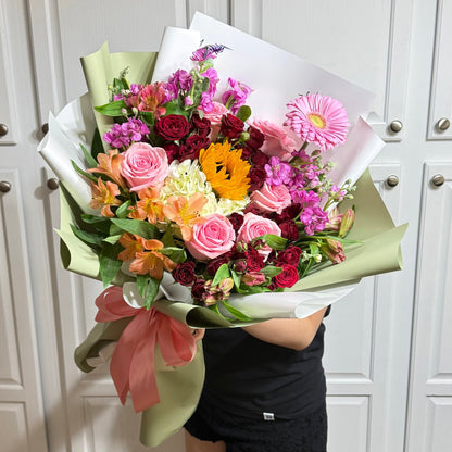 Colorful bouquet with pink roses, a sunflower, and mixed flowers wrapped in green paper.