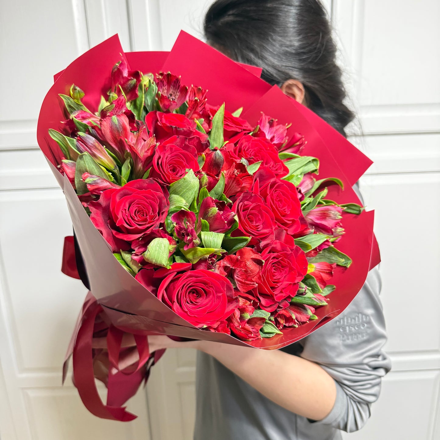 A person holds a large bouquet of red roses and alstroemeria wrapped in red paper.