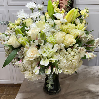 Large white and light yellow floral arrangement in a dark vase on a counter.
