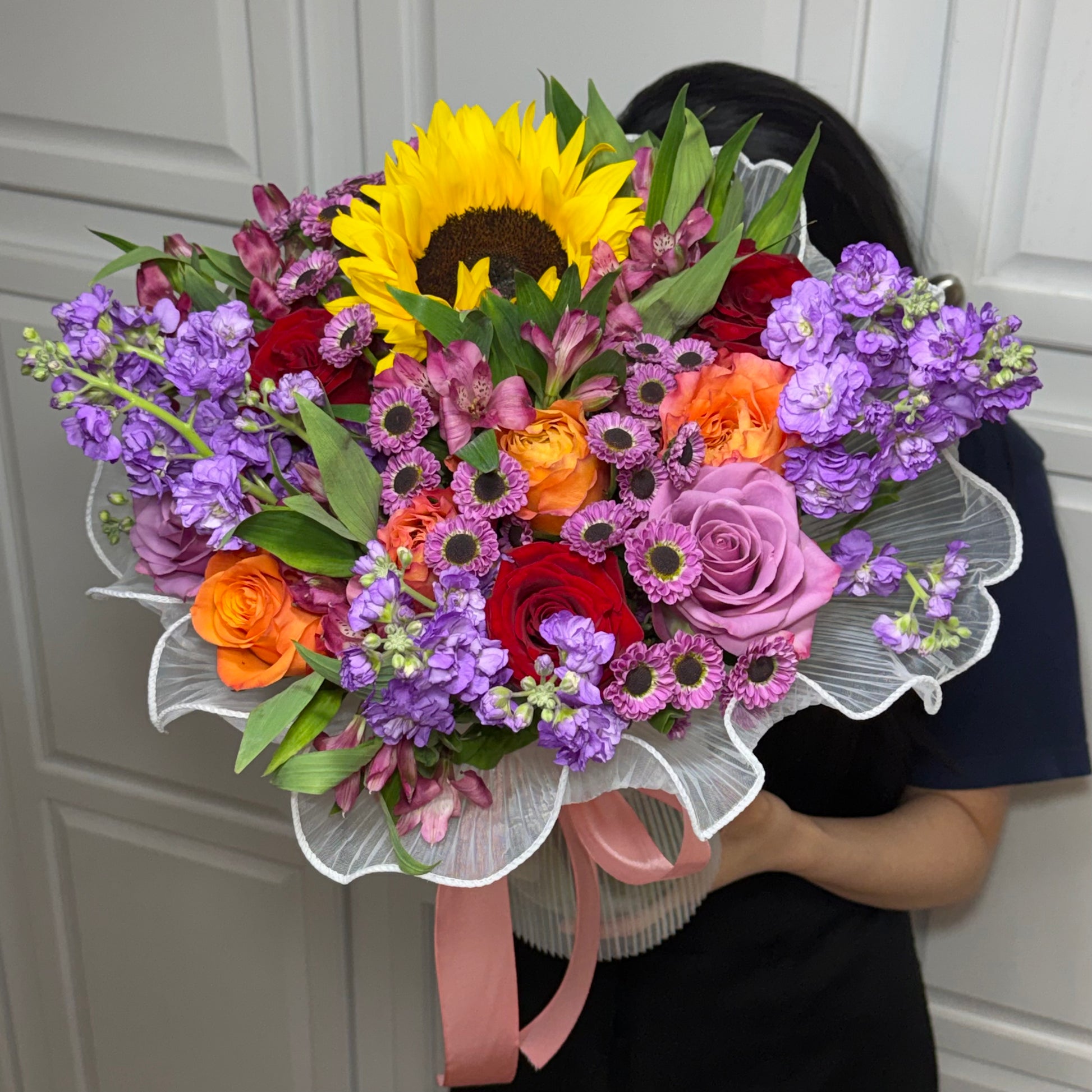 A vibrant mixed flower bouquet with a sunflower, roses, and purple blooms.