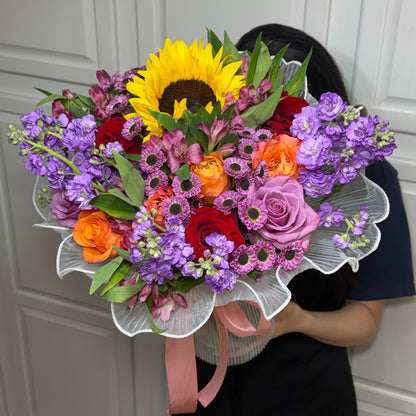 A vibrant mixed flower bouquet with a sunflower, roses, and purple blooms.