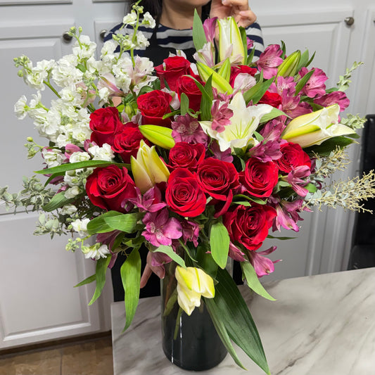A large, vibrant bouquet of red roses, white lilies, and purple flowers in a dark vase.